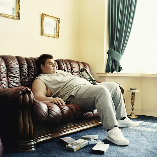 Young Man Lies Slouched on a Sofa Watching Videos and Holding a Packet of Crisps