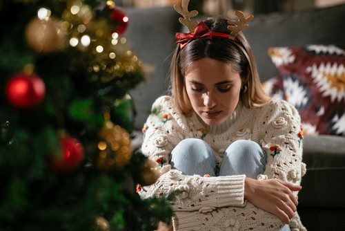 Young sad woman sitting near the Christmas tree. Depressed woman during Christmas holiday at home.