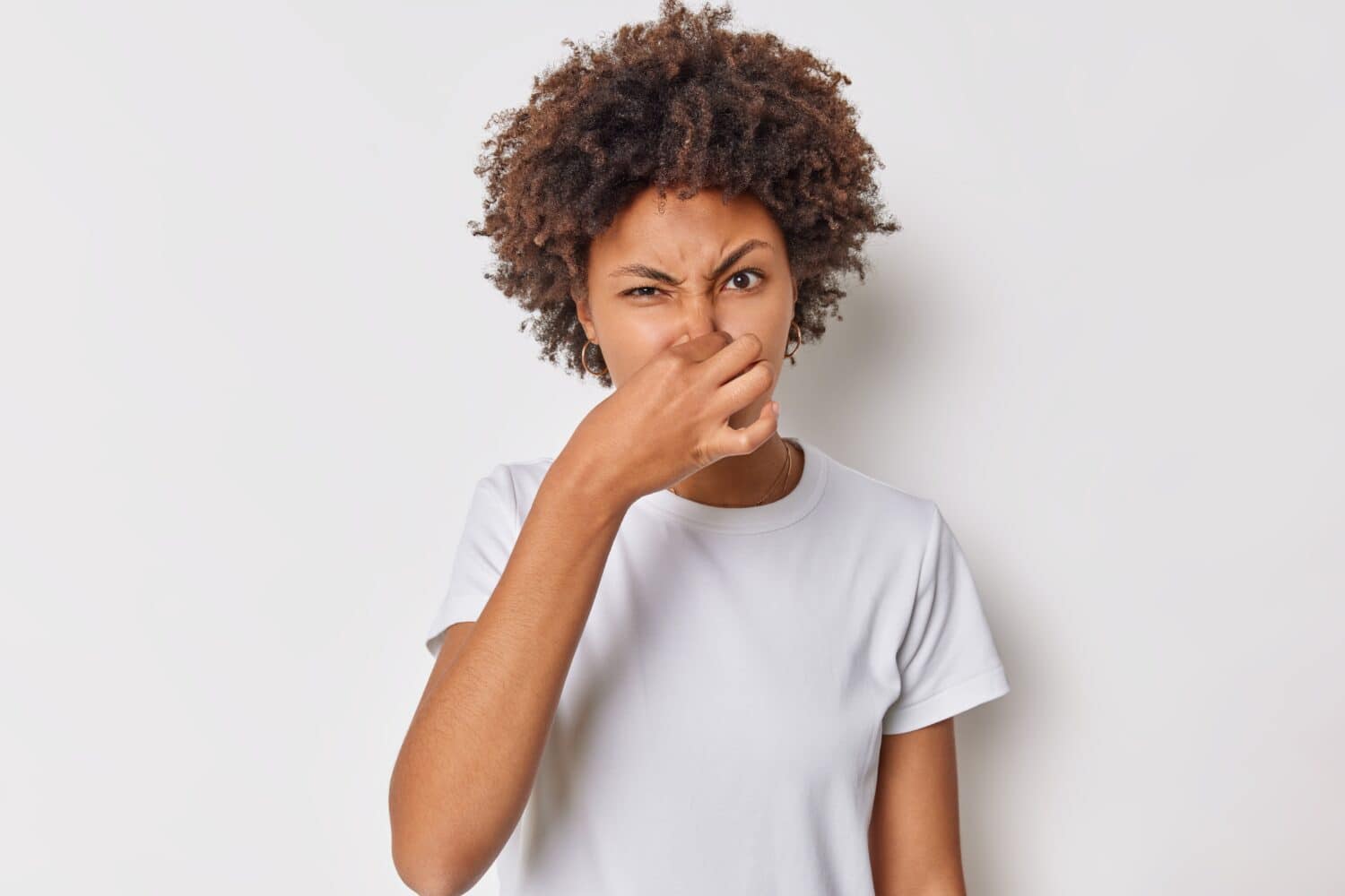 Fie something stinks. Displeased curly haired woman covers nose smells something unpleasant has disgusting expression wears casual t shirt isolated over white background. Bad smell coming from kitchen