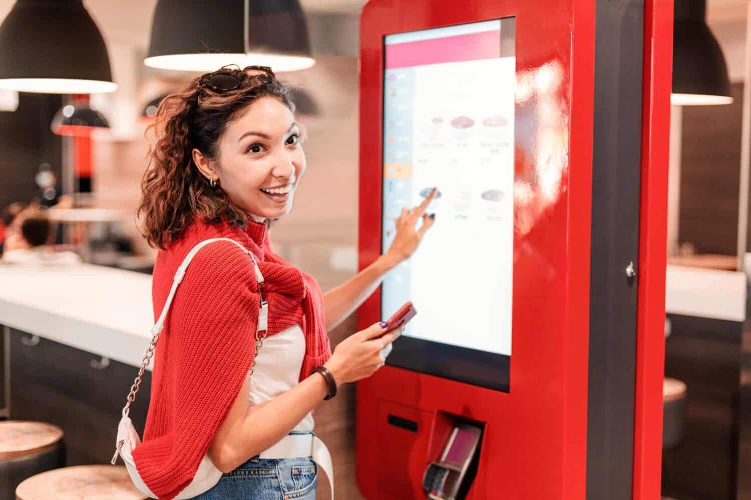 A girl orders food and lunch at a fast food restaurant using a self-service kiosk or a terminal with a screen. Modern commerce equipment