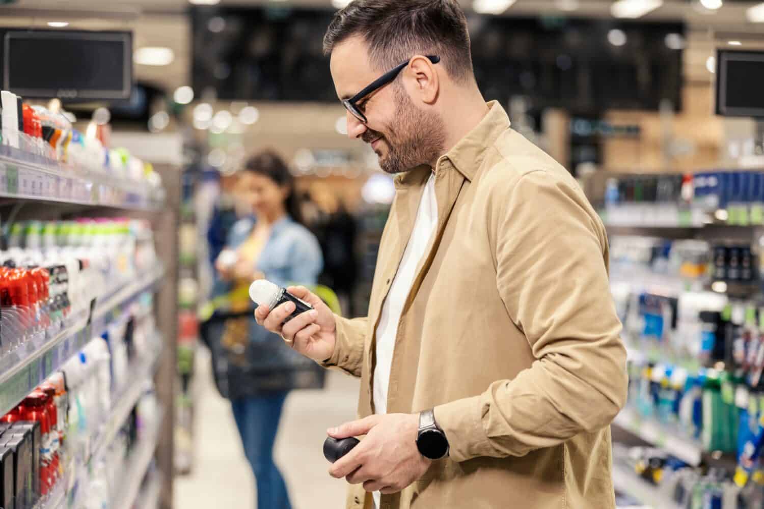 A happy man is standing next to a shelves in with deodorants in supermarket and choosing one.