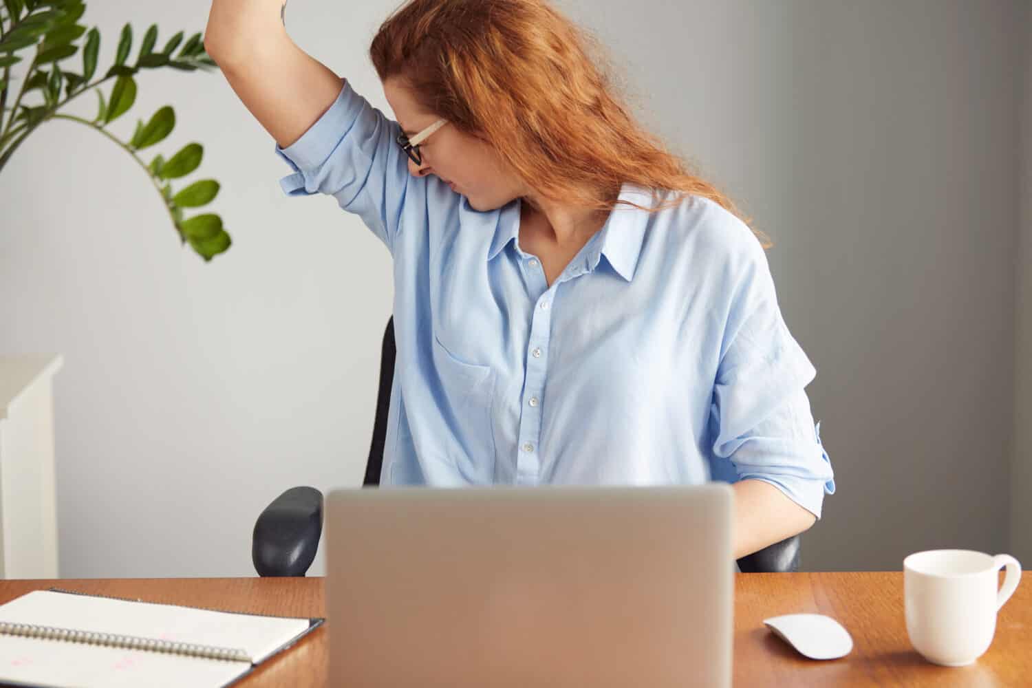 Portrait of a young woman wearing blue shirt disgusting with bad smell of her wet armpit while working in the office. Something stinks, negative human emotions, facial expressions, feeling reaction