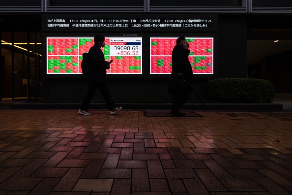 Two silhouetted individuals walk along a wet brick sidewalk at night, illuminated by a brightly lit large digital stock market display board showing red and green squares of stock information. The display shows a main index value of 
