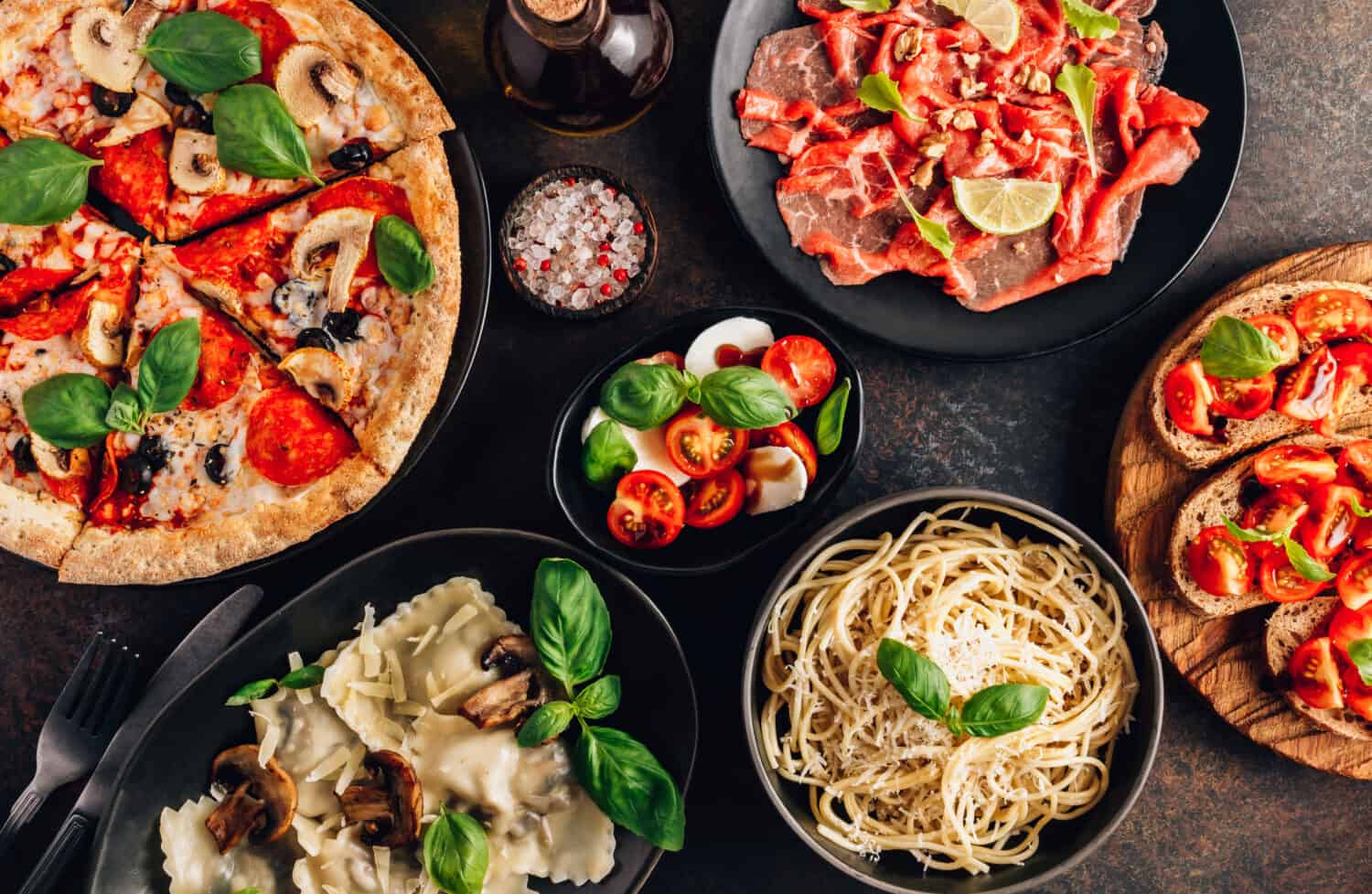 Full table of italian meals on plates Pizza, pasta, ravioli, carpaccio. caprese salad and tomato bruschetta on black background. Top view