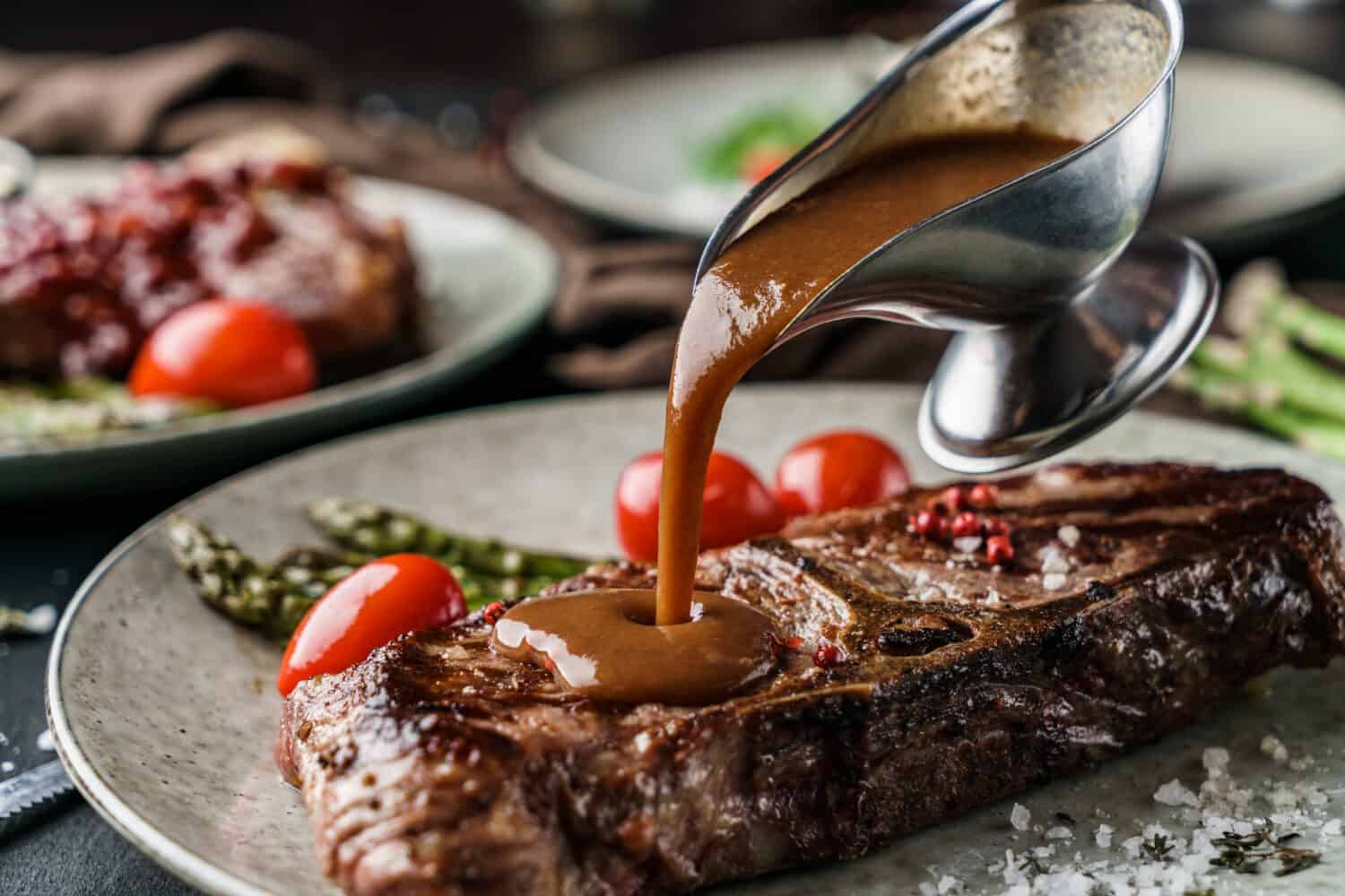 Delicious grilled beef steak with spices on plate with tomatoes and barbecue sauce over dark background. Concept of bbq food, close up