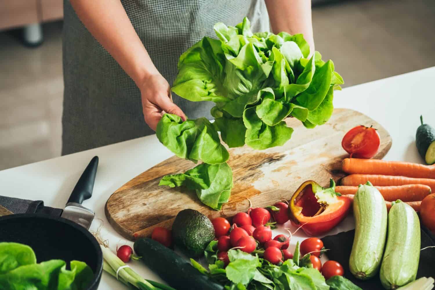 Close Up Photo of Woman Hands Making Fresh Salad on a Table Full with Organic Vegetables. An anonymous housewife making lunch with fresh colorful vegetables at kitchen table.