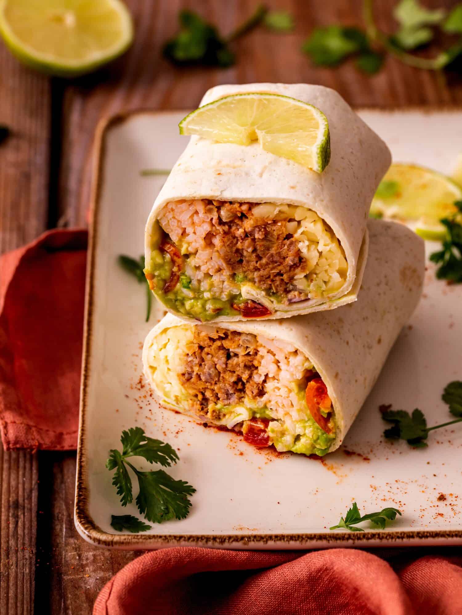 Mission burrito with refried beans, beef sauce, guacamole dip, red rice, and shredded cheese rolled in a tortilla and served with sour cream dip and lime juice on a square plate on a wooden table