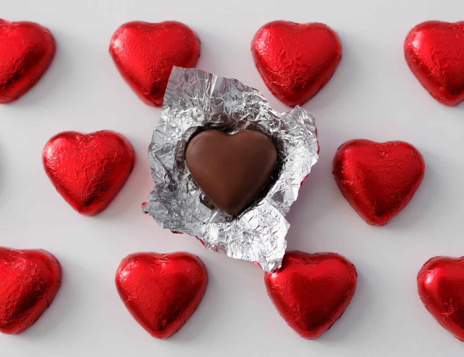 Love and relationship concept - close up of wrapped and unwrapped heart shape chocolate candies in red foil over white background