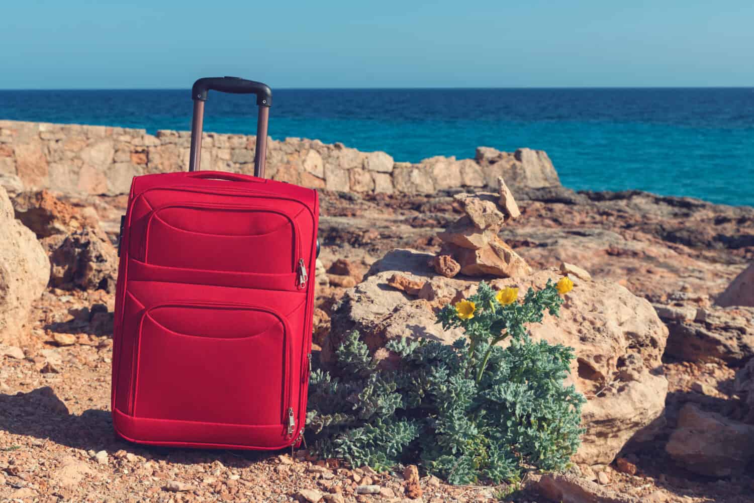 Red suitcase and yellow horned poppy on a rocky sea shore in Cap de Salines in Majorca. Selective focus