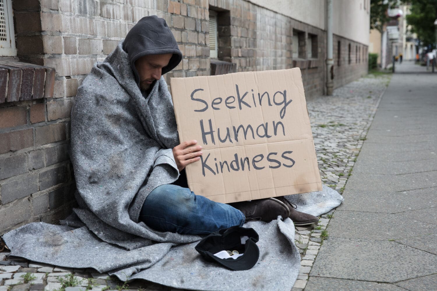 Male Beggar In Hood Showing Seeking Human Kindness Sign On Cardboard