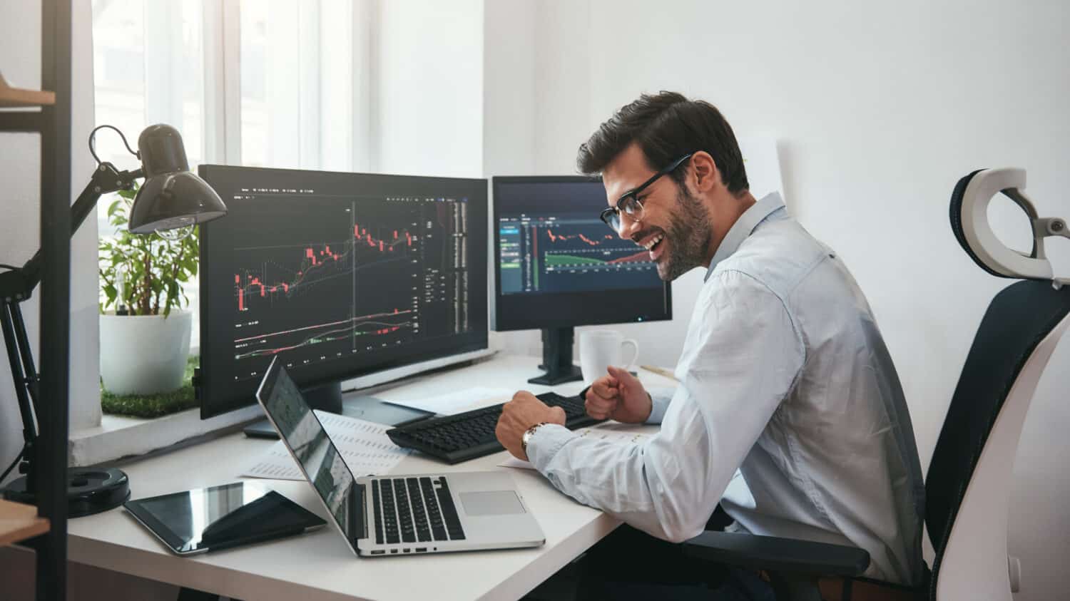 Lucky day. Happy young businessman or trader in formalwear and eyeglasses using laptop and smiling while sitting in his modern office