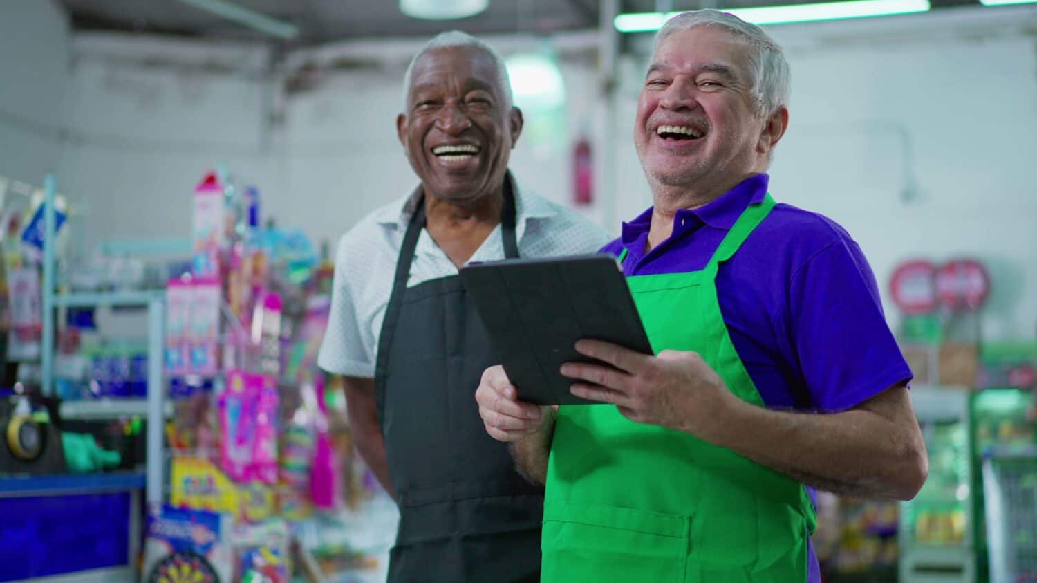 Joyful diverse Brazilian senior staff workers of supermarket chain smiling at camera with table and uniforms. African American older employee and a caucasian person laughing together