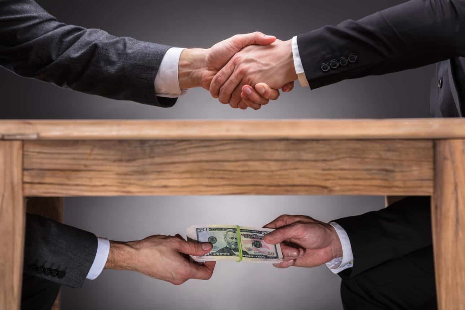 Close-up Of Two Businesspeople Shaking Hand And Taking Bribe Under Wooden Table On Grey Background