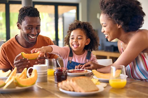 Family Shot With Parents And Daughter At Home Having Breakfast Spreading Jam On Bread At Table