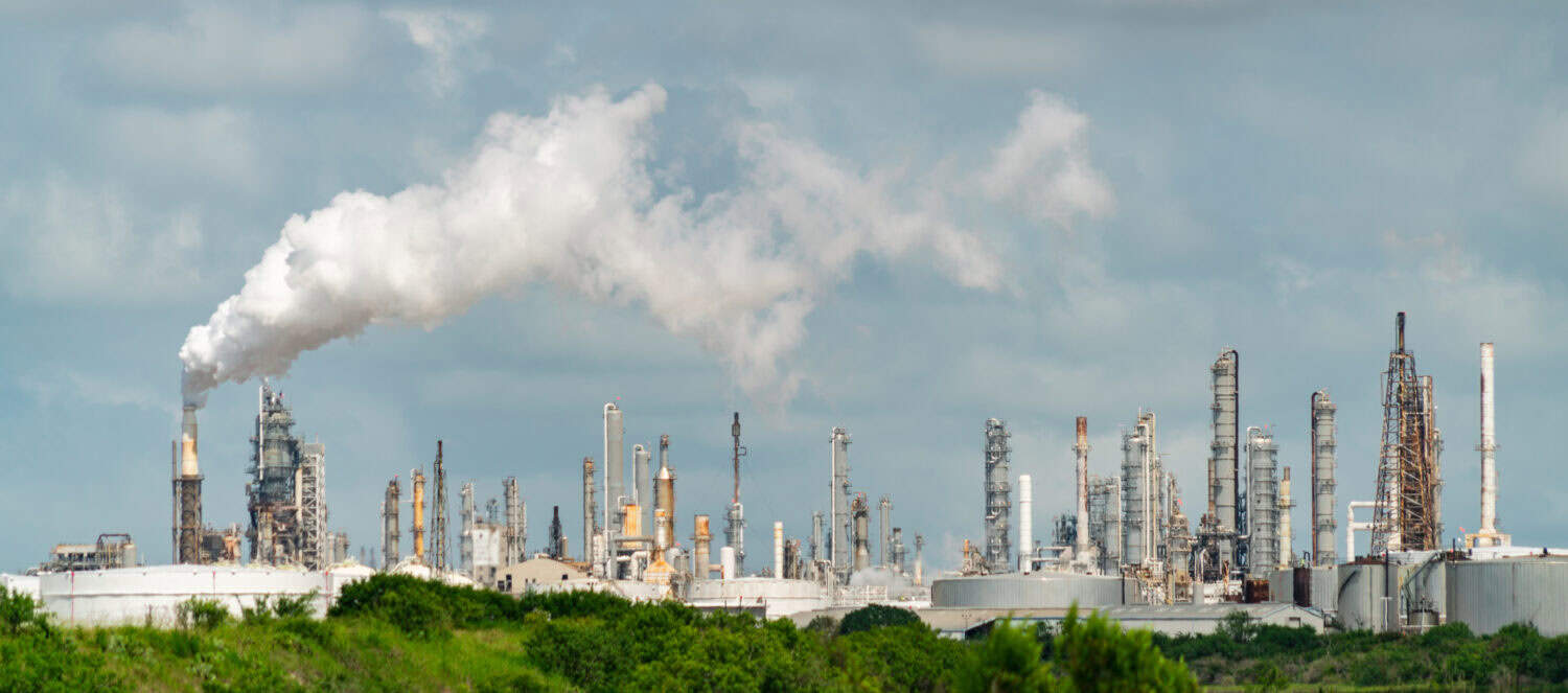 green trees in front of dirty petrochemical factory or oil refinery polluting carbon and pollution into the cloudy smog skies cuasing climate change and global warming and extreme weather events