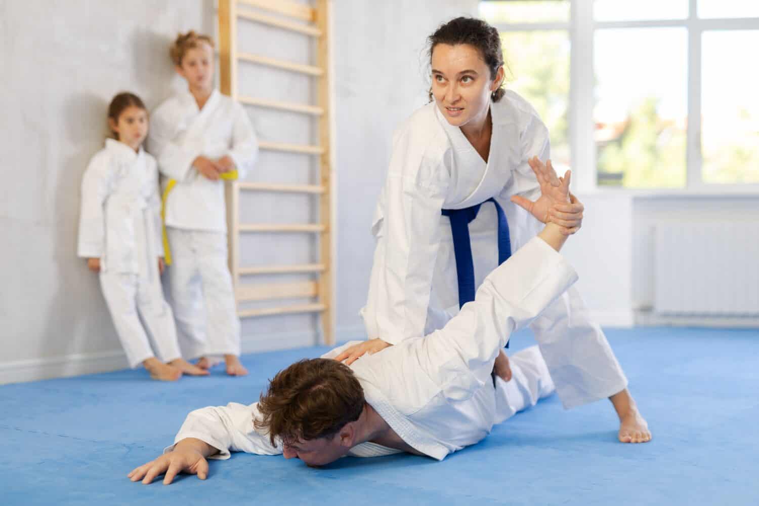 Concentrated young woman attending family karate class, applying armlock technique to husband while preteen children in kimonos watching training sparring in background