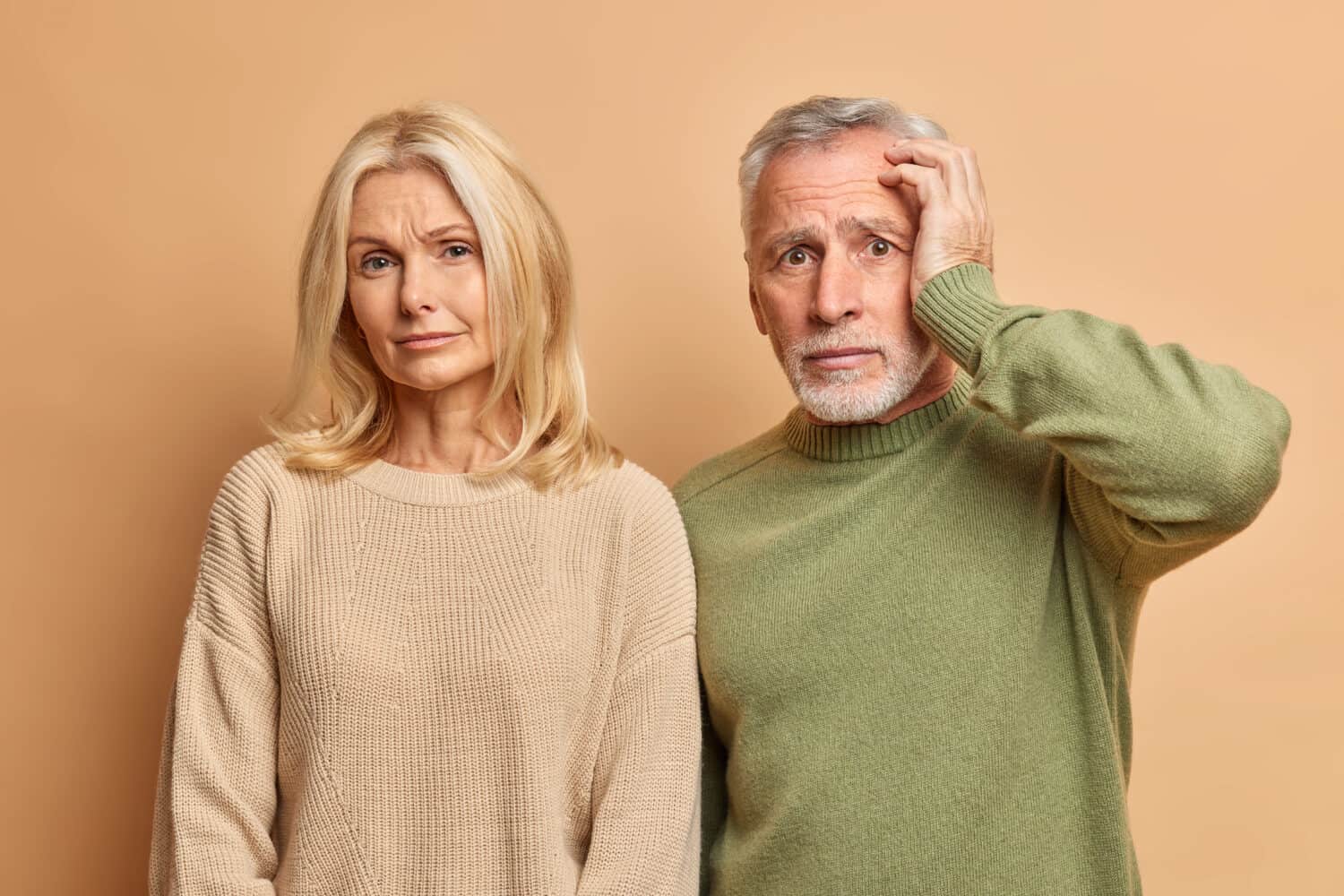 Tired displeased blonde wrinkled woman looks calmly at camera and embarrassed troublesome man looks with panic and fearful expression isolated over brown wall. Old people emotions reaction concept