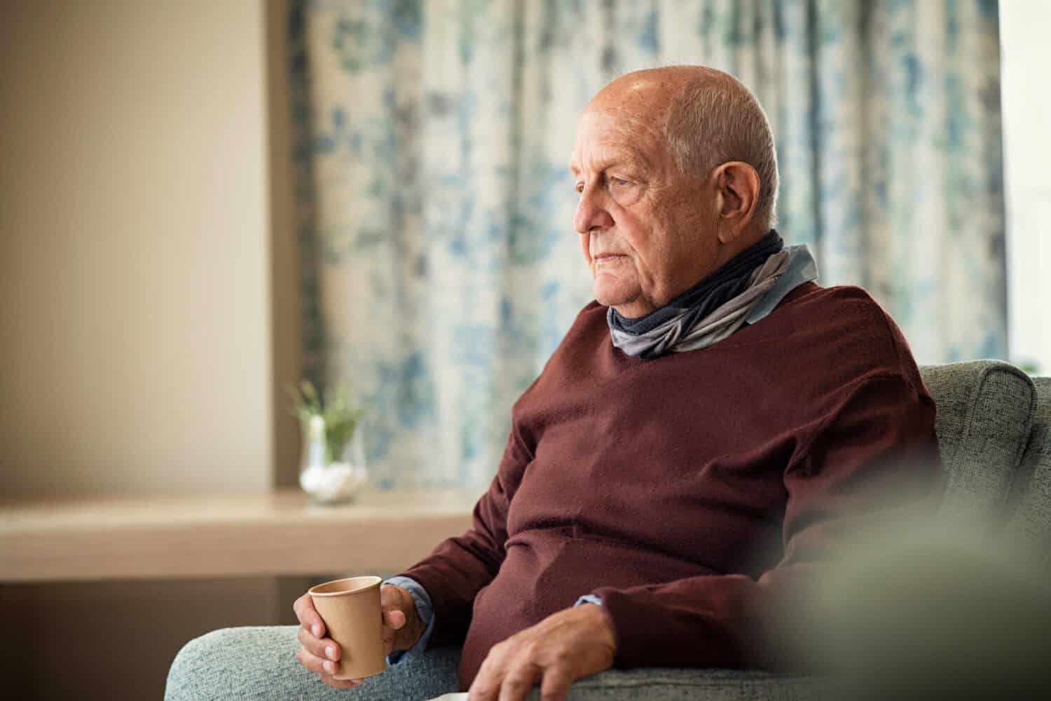 Depressed senior man sitting on armchair holding disposable cup of coffee and thinking. Frustrated retired man sitting on sofa. Sad mature man sitting alone at nursing home with sad expression.