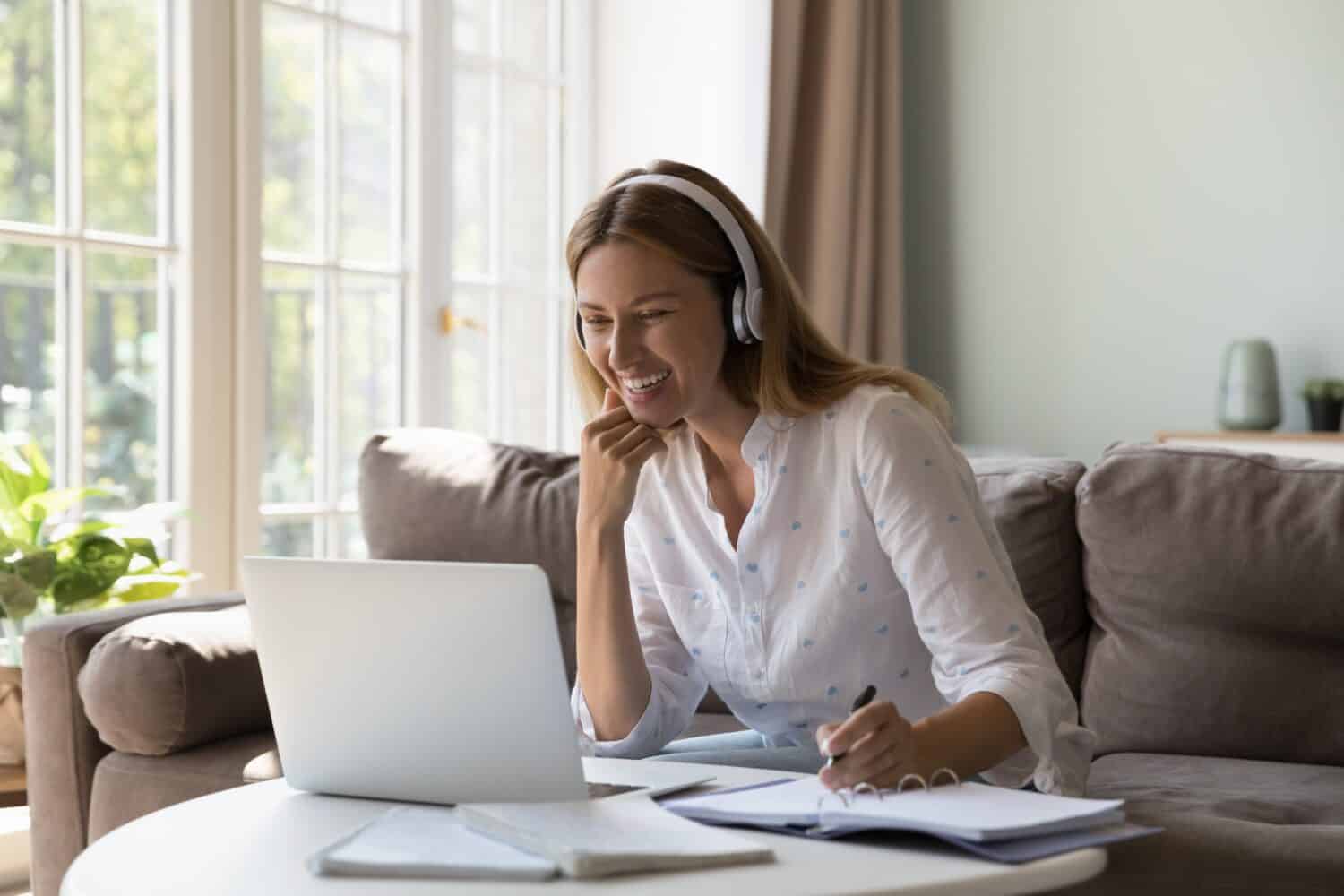 Woman in headphones studying from home using video call app, sit at table with laptop, makes notes improve English knowledge with on-line tutor. Remote class, new skills, virtual meeting event