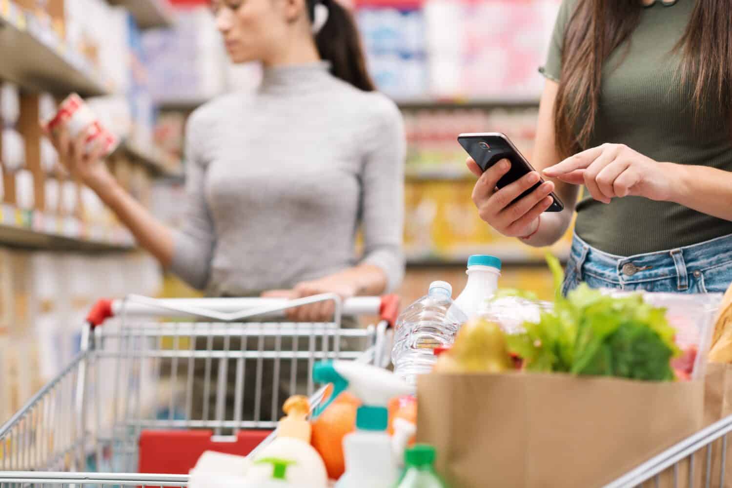 Women doing grocery shopping together at the supermarket, one is using her smartphone in the foreground