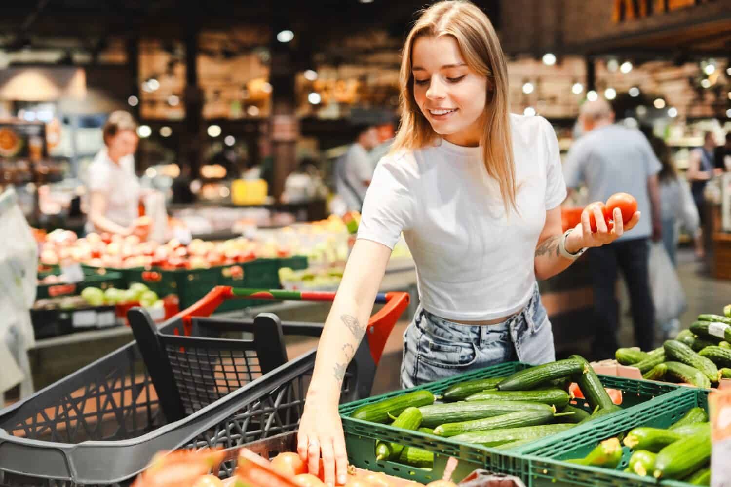 Young woman happily shops for fresh produce in a grocery store, selecting tomatoes and cucumbers for a healthy lifestyle. The vibrant vegetables stand out against a blurred background of shoppers
