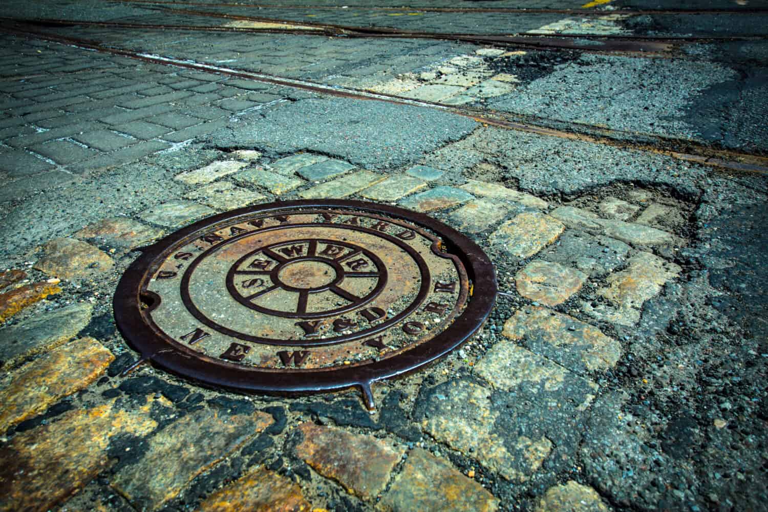 Manhole drain cover on rough old cobblestone and asphalt street at historic Brooklyn Navy Yard