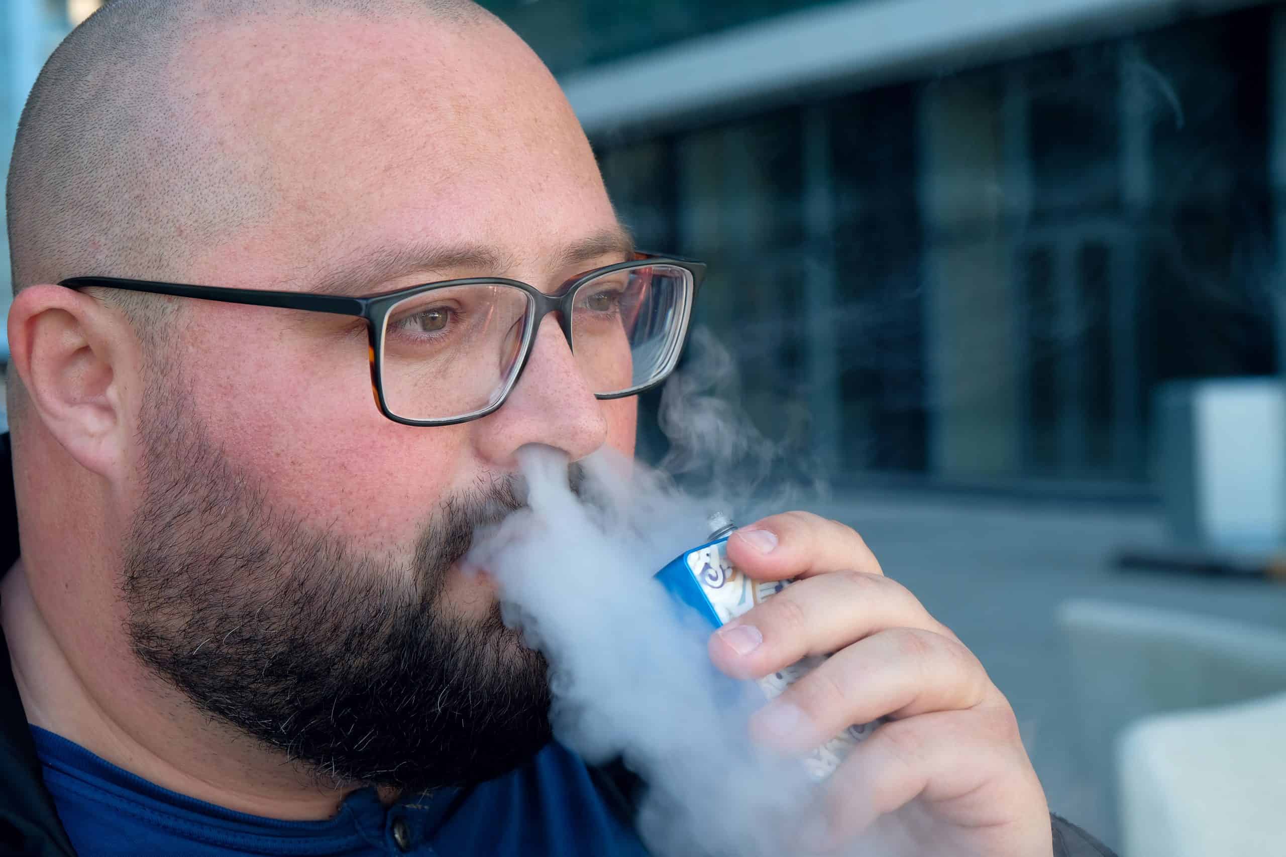 A young man releases cigarette smoke through his nose and mouth. A bald man takes a puff, smokes an electronic cigarette. The concept of drug addiction, unhealthy lifestyle, bad habits, wireless technologies. Nicotine addiction. Portrait, close-up.