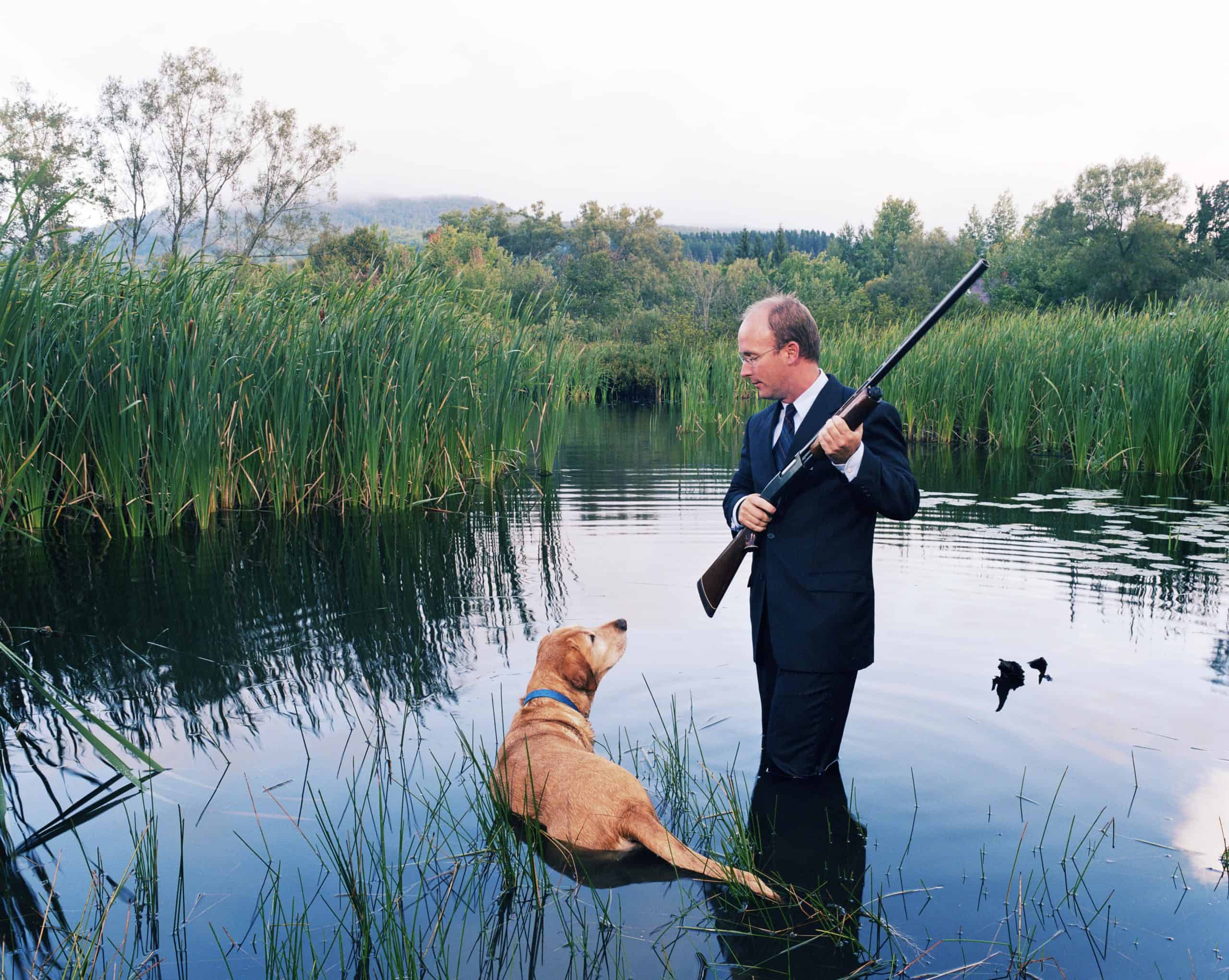 gun Vermont | Businessman knee deep in water holding shotgun, looking at dog