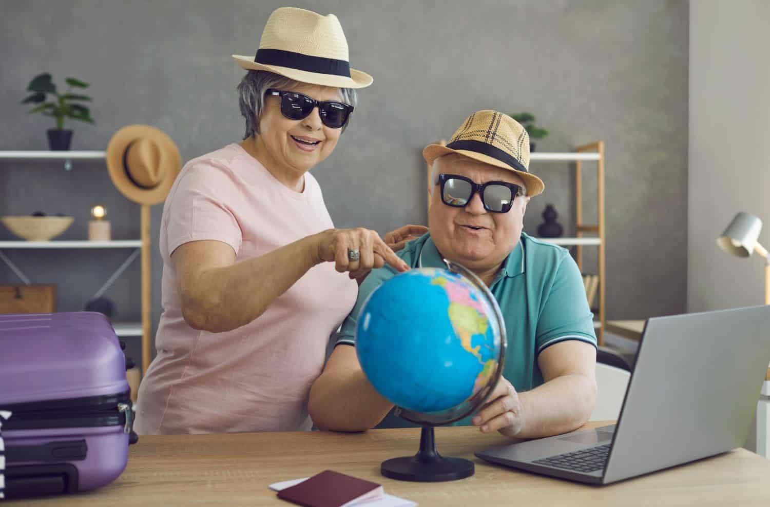 Happy excited retired senior couple planning holiday trip together. Cheerful old people in hats and sunglasses choosing their dream destination on Earth globe. Traveling and active vacation concept