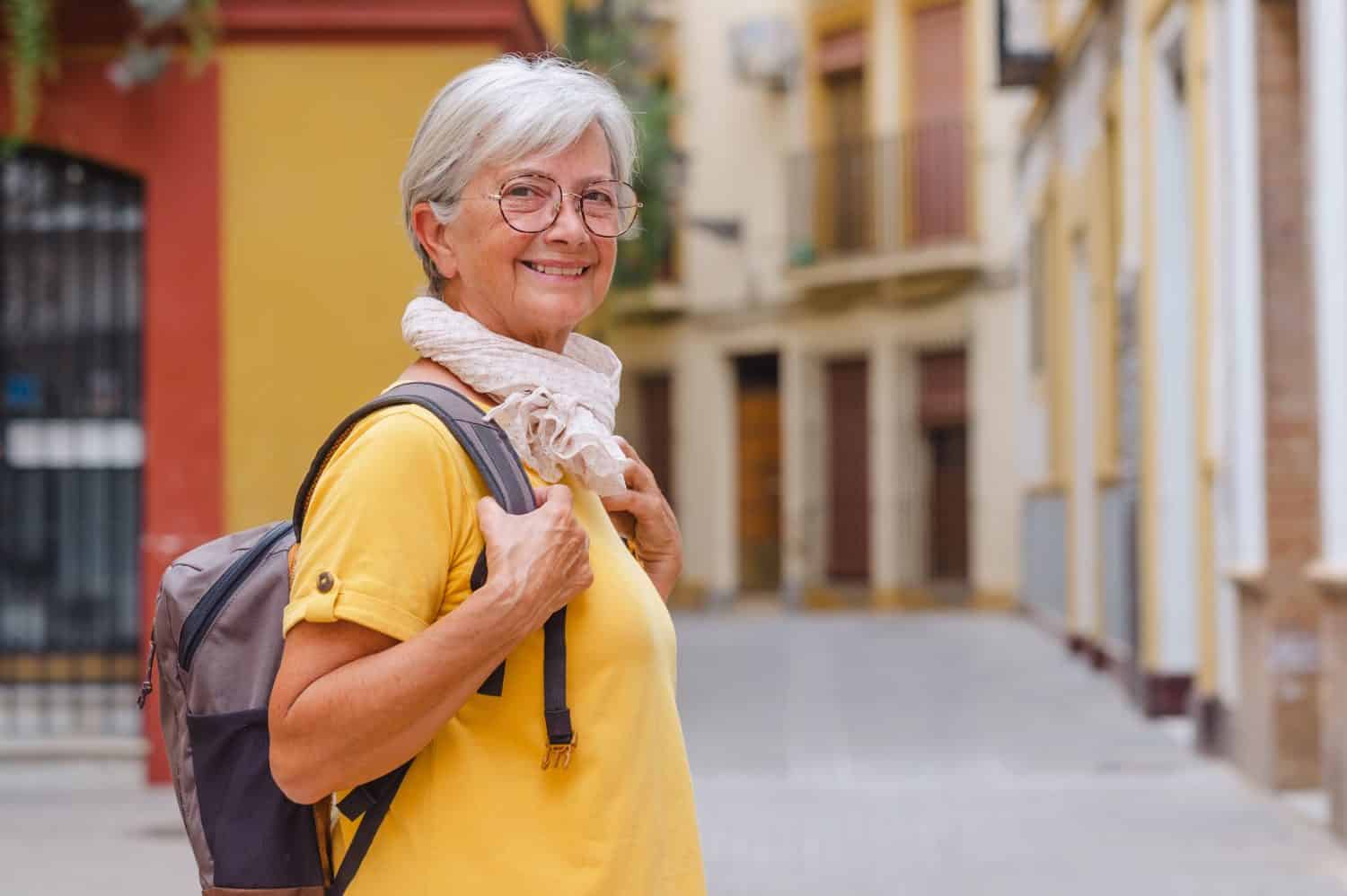 Portrait of senior traveler woman carrying backpack visiting the old town of Seville, smiling elderly lady enjoying travel and discovery