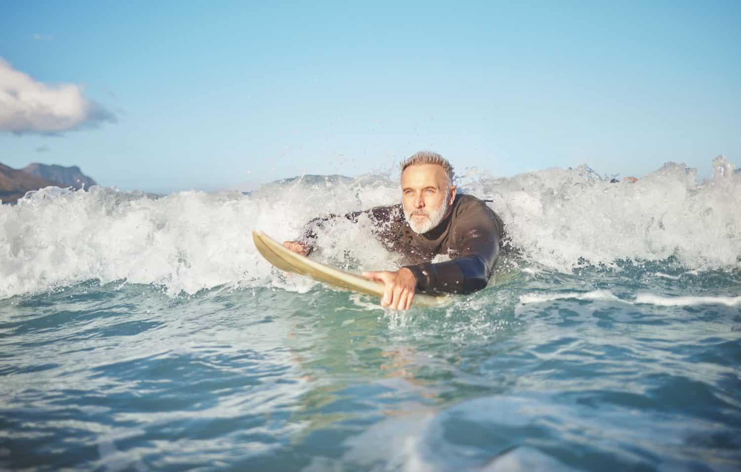 Ocean waves, senior man surfing on beach and healthy fitness lifestyle in Australia summer holiday. Elderly surfer swimming with surfboard, sea water exercise and relax in retirement travel vacation