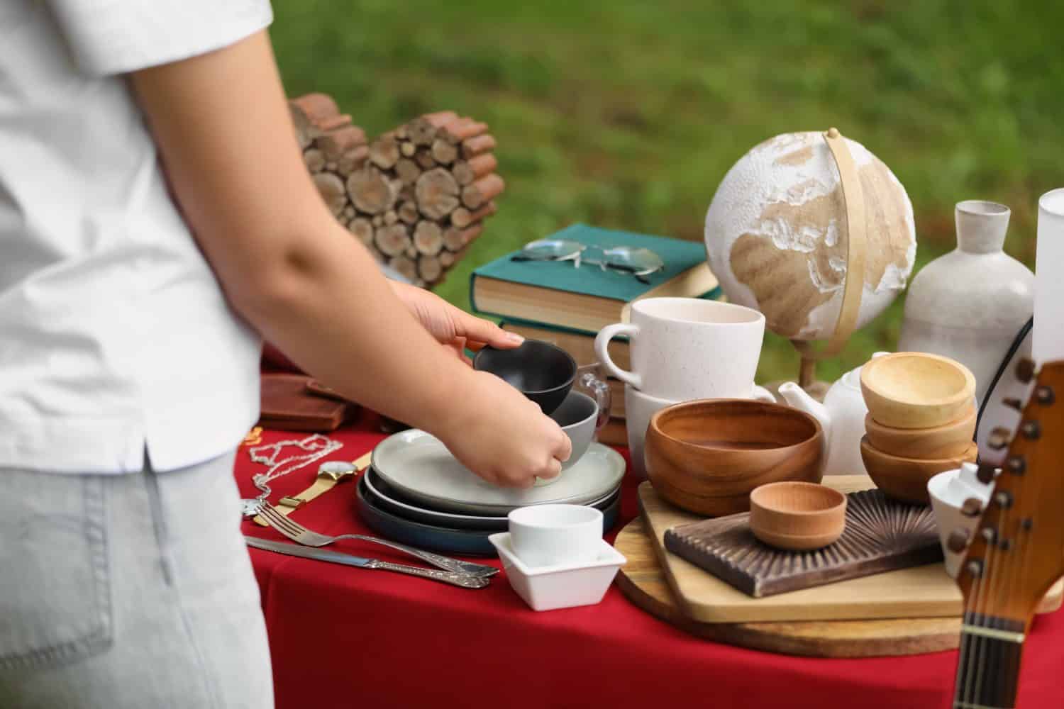 Woman holding beautiful bowls near table with different items on garage sale, closeup