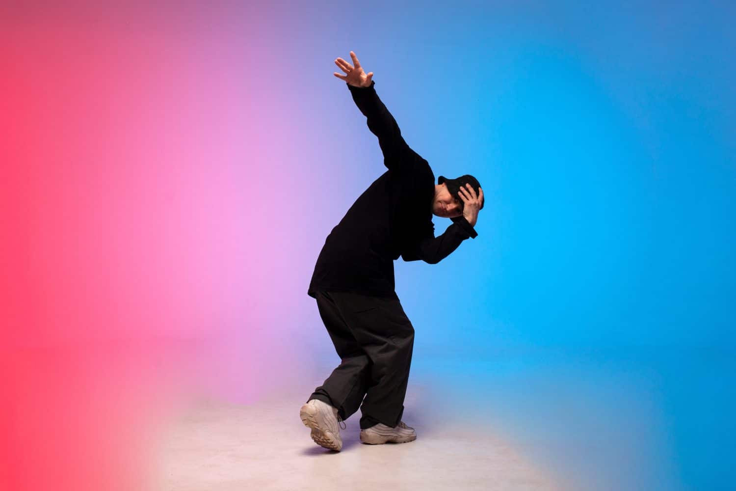 young guy dancer in black clothes break dancing in neon lighting, man performs hip hop movement in red blue club lighting