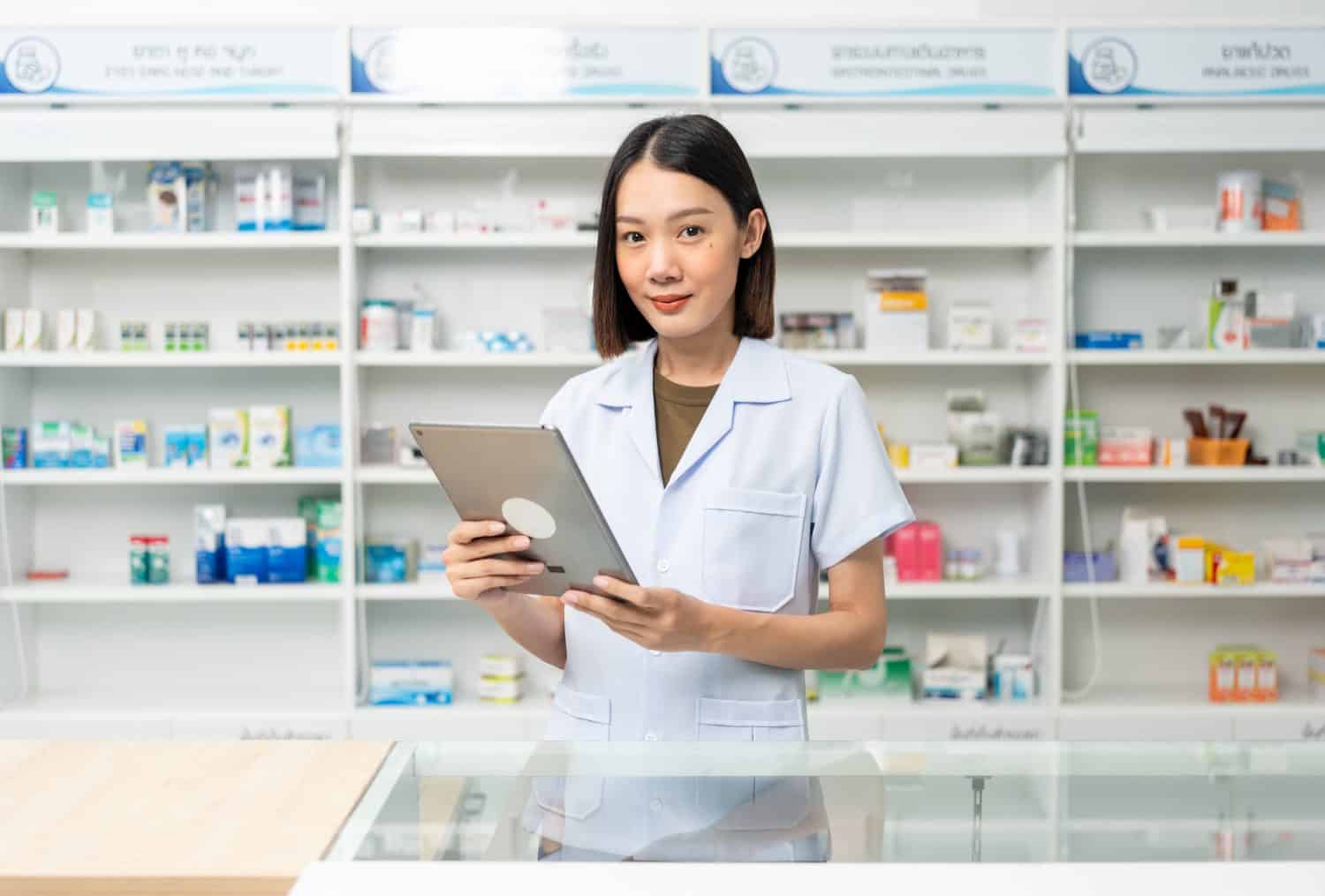 Beautiful asian woman pharmacist checks inventory of medicine in pharmacy drugstore. Professional Female Pharmacist wearing uniform standing near drugs shelves working with tablet.