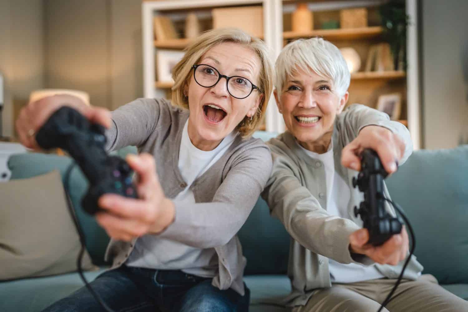 Two senior women caucasian friends or sisters happy old siblings pensioner playing video game console using joystick or controllers while sitting at home real people family leisure concept copy space