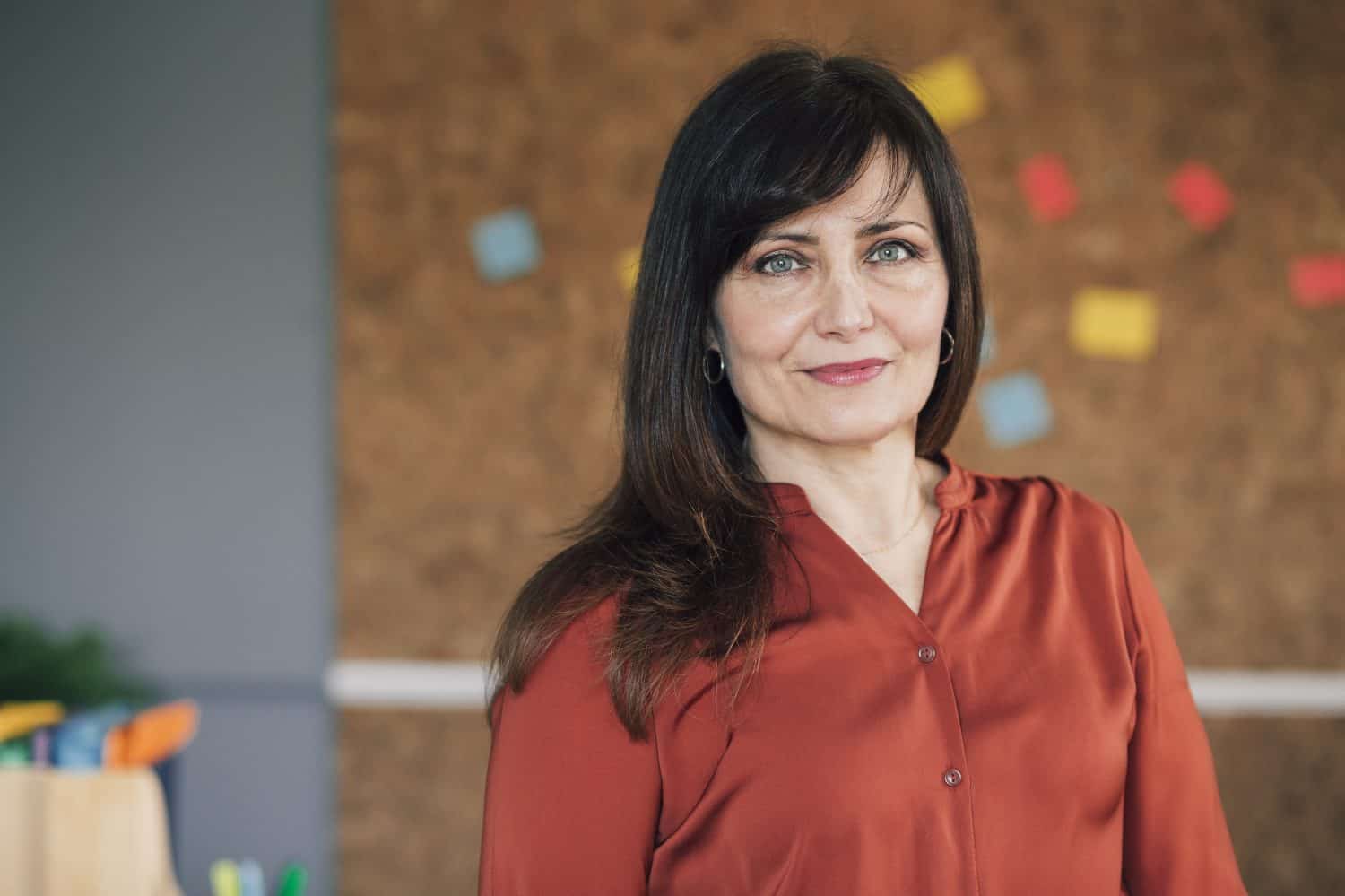 Portrait of a mature, female business owner in her office. She is standing in front of a cork board and is smartly dressed, smiling for the camera.