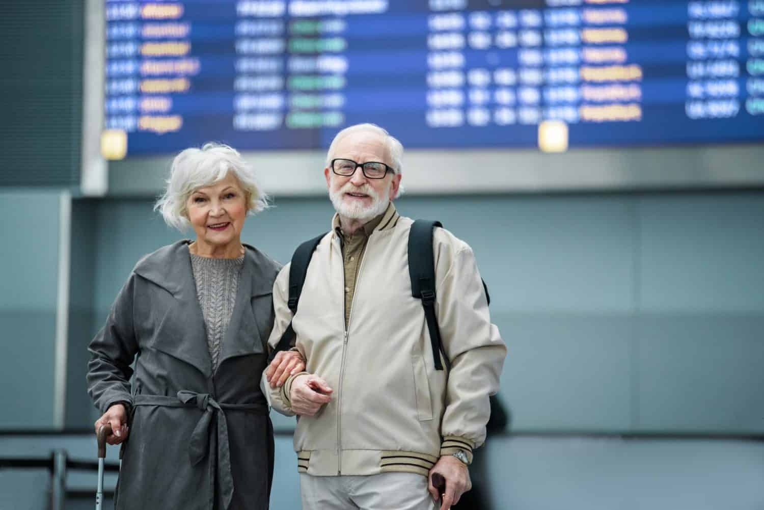 Travel abroad. Portrait of elegant aged man and woman are standing together against timetable at international airport and looking at camera with joy. Copy space in the right side