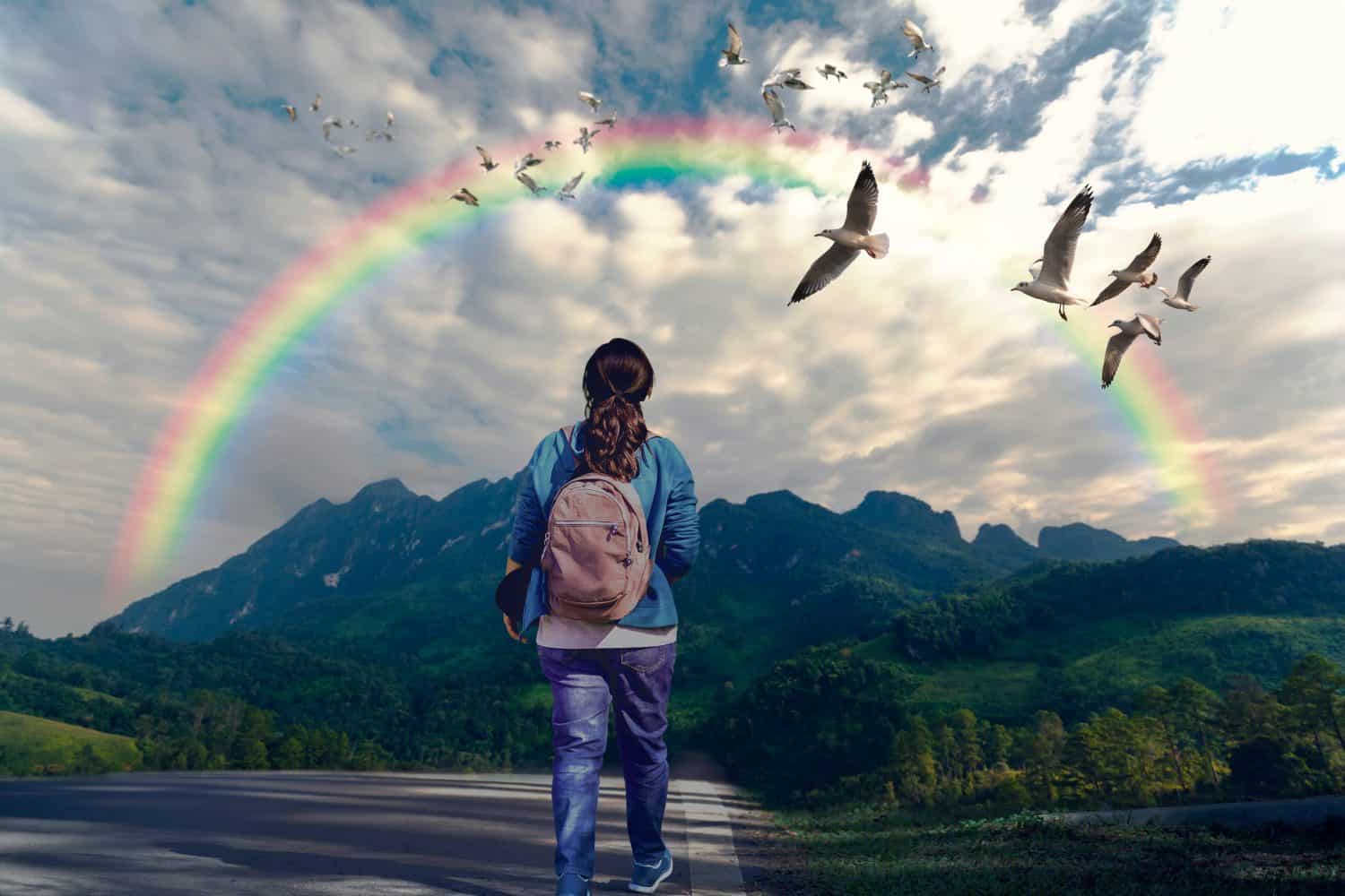 Follow your dream concept. A woman walking along the path to mountain and looking rainbow in the sky.