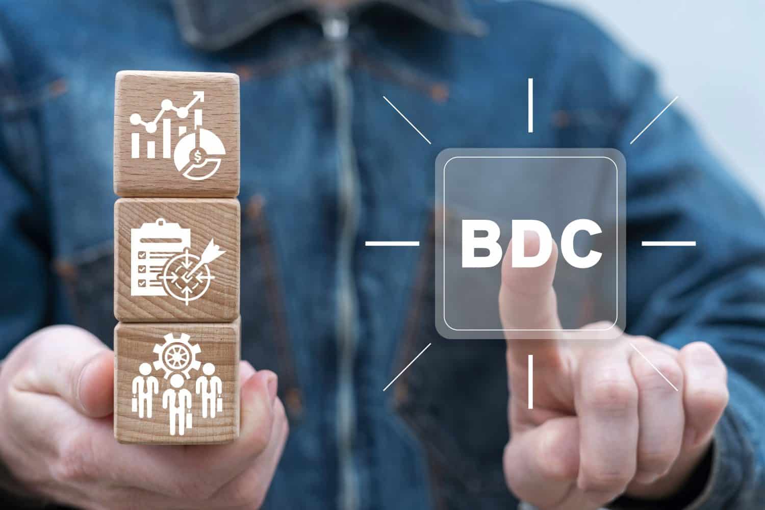 Businessman holding wooden blocks with icons and sees virtual button with abbreviation: BDC. Concept of BDC Business Development Company.