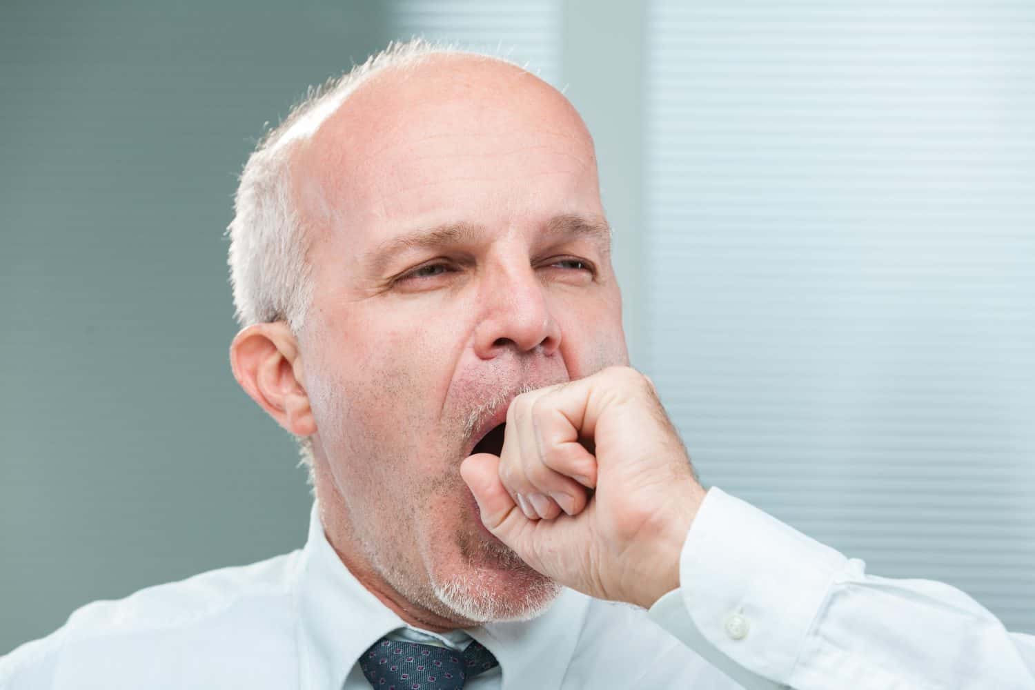 Aged clerk yawns, covering his mouth ineffectively. The close-up reveals a man tired, perhaps bored, needing oxygen or hinting at a transition. Dressed as a senior businessman in shirt and tie