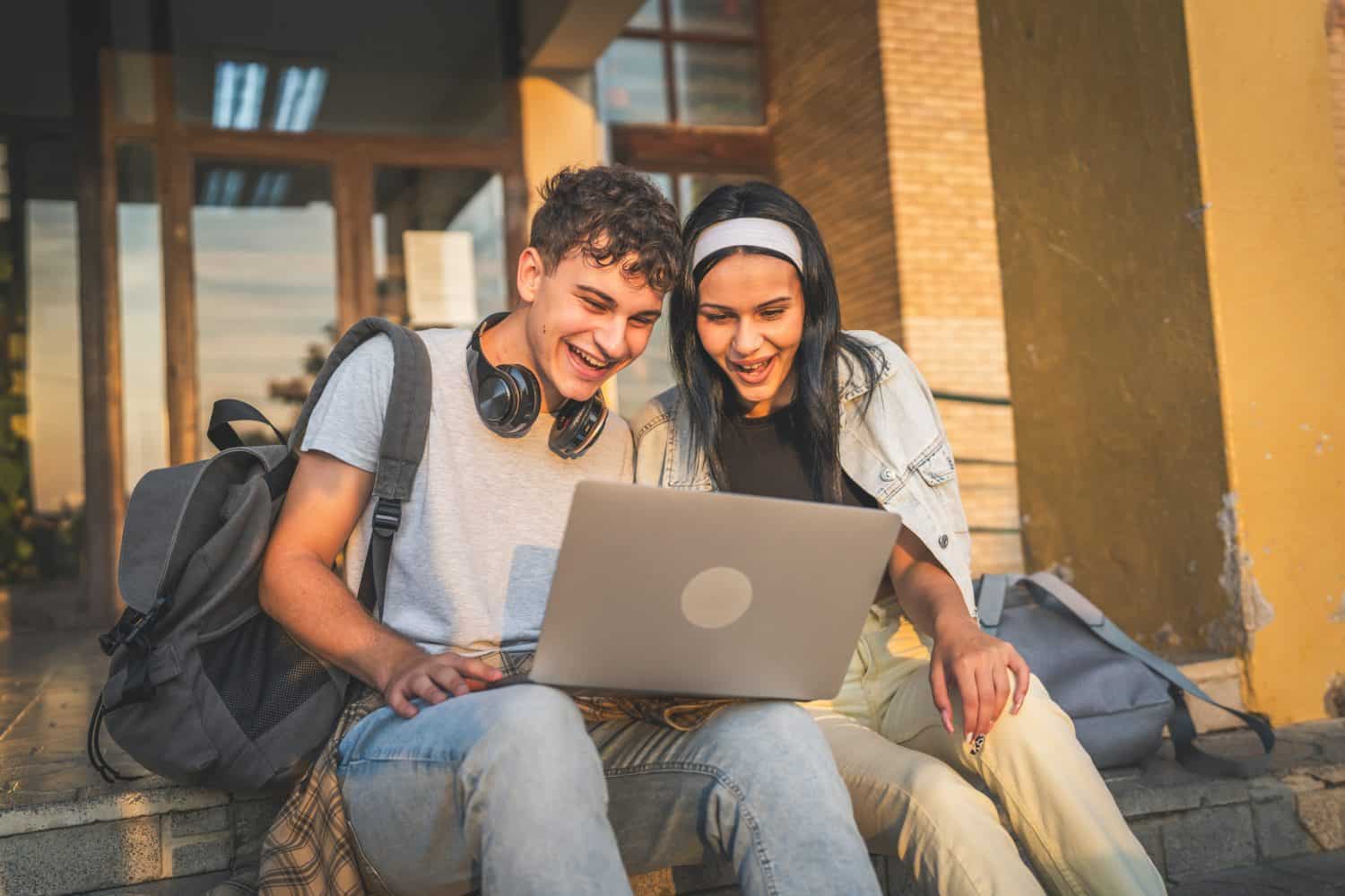 teenage couple caucasian male and female boyfriend and girlfriend students man and woman watch video movie on laptop computer at school while sit outdoor in sunny day happy smile have fun online