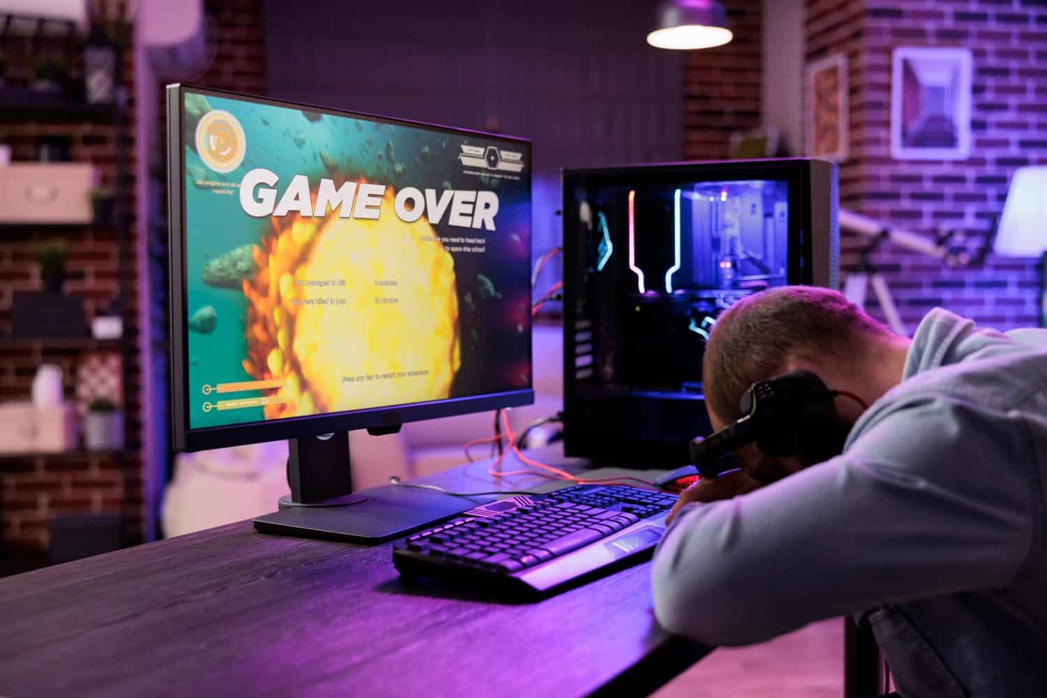 Gamer feeling gutted after losing singleplayer action videogame level, being outsmarted by enemies. Uspet man placing head on desk in frustration after seeing game over screen