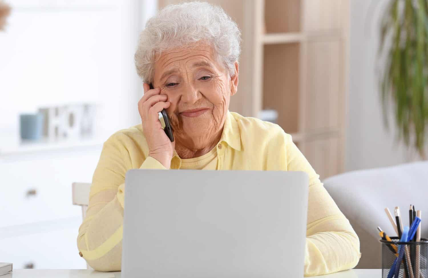 Senior woman with laptop talking by mobile phone at home