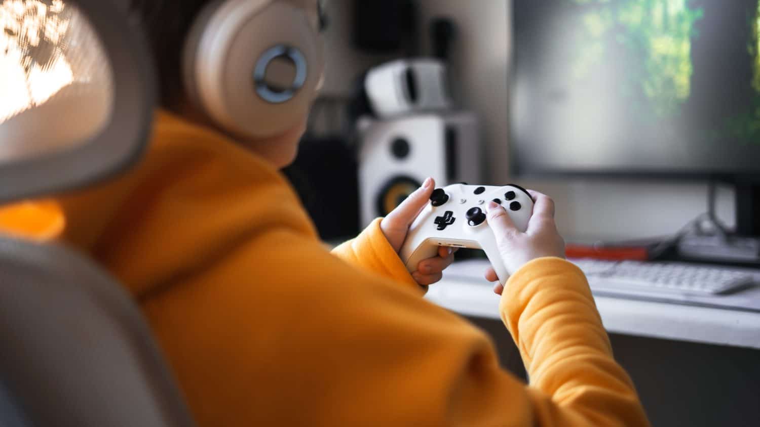 Teenage girl with headset is sitting in a comfortable computer chair, holding a white gamepad in her hands and playing a game on a PC or console.