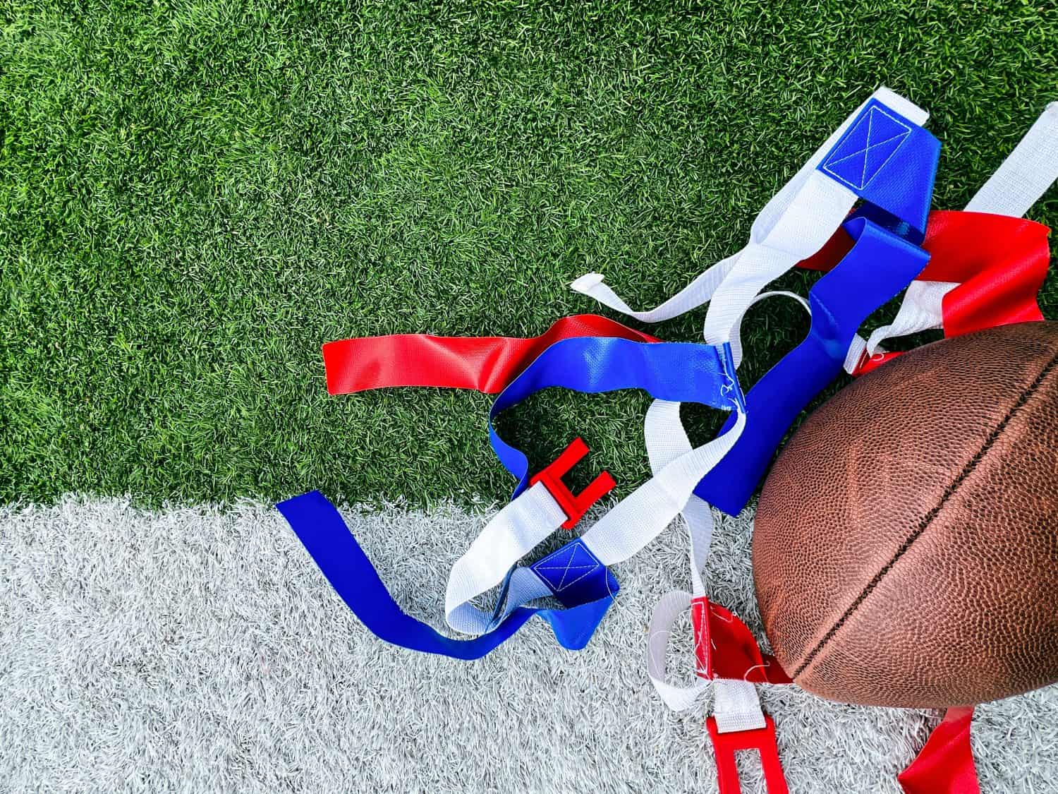 Flag Football Equipment on Green Turf. White belts with red and blue flags used for playing flag football. A football lays to the side. Set against the white and green turf of a football field.