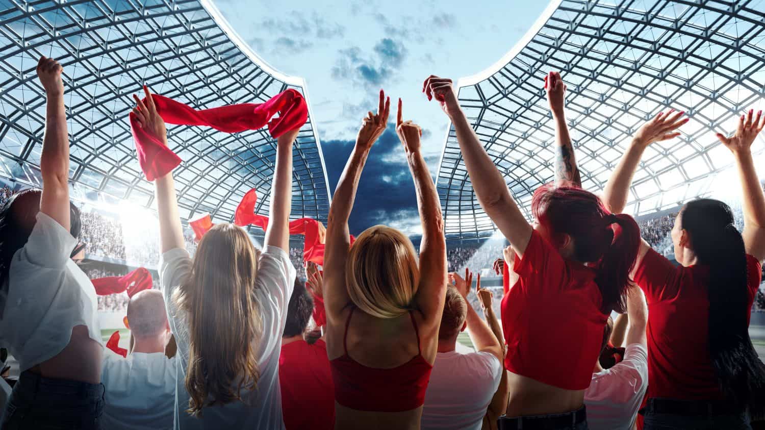 Vibrant crowd of enthusiastic sports fans raises their arms and waves red scarves in celebration against modern stadium with clear sky above. Concept of support, international match, betting, games.