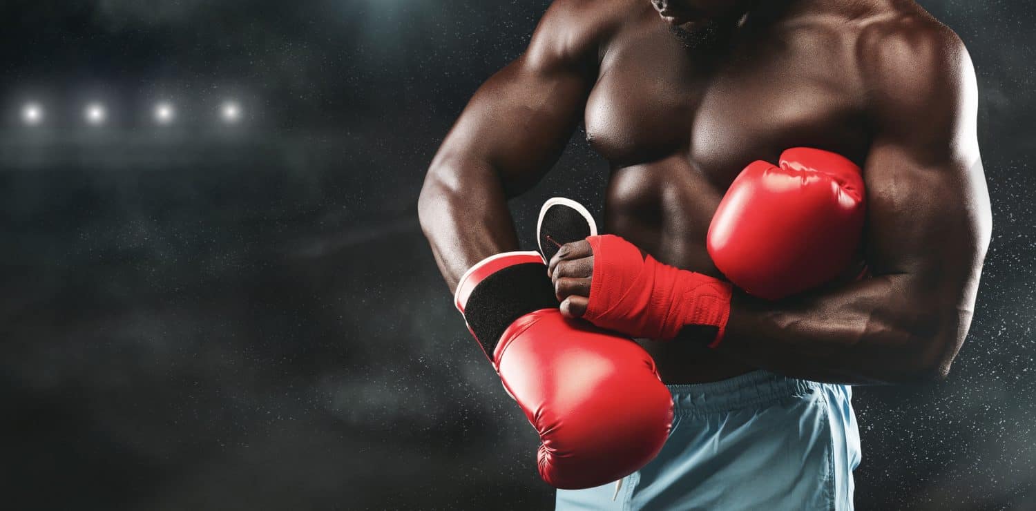 A muscular boxer adjusts his red boxing gloves in a dimly lit boxing ring, ready for a fight.