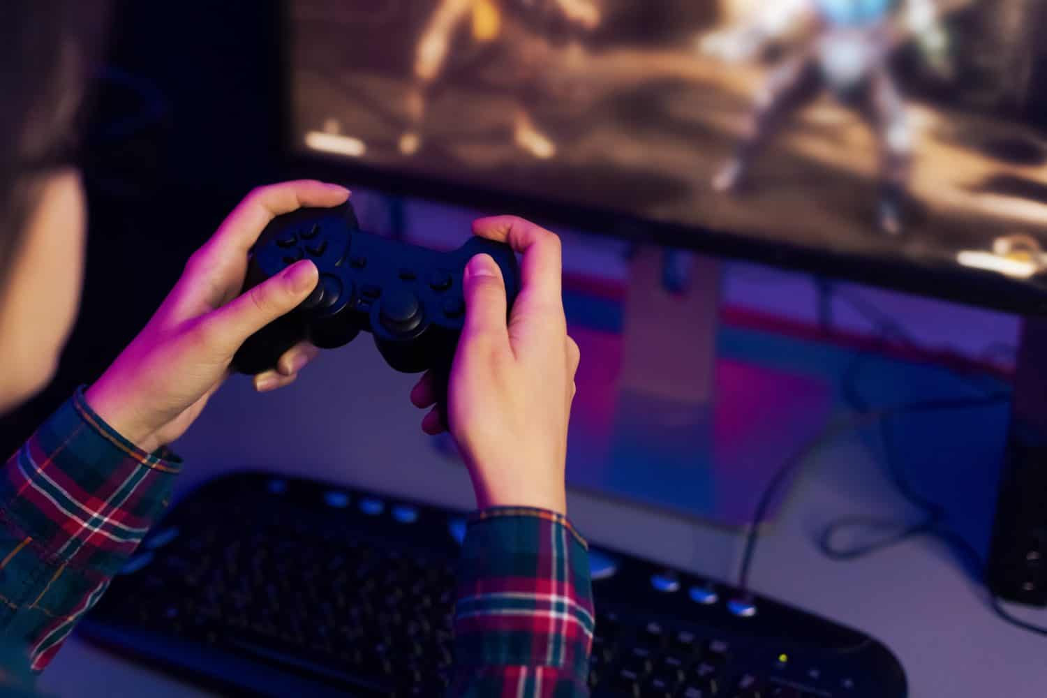 A persons hands hold a black video game controller, with their fingers resting on the buttons, playing a video game on a computer monitor, which is lit up with a colorful screen displaying the action.