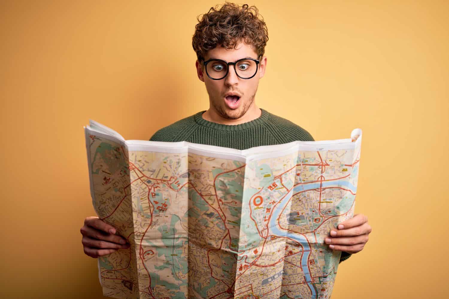 Young blond tourist man on vacation with curly hair holding city map over yellow background scared in shock with a surprise face, afraid and excited with fear expression