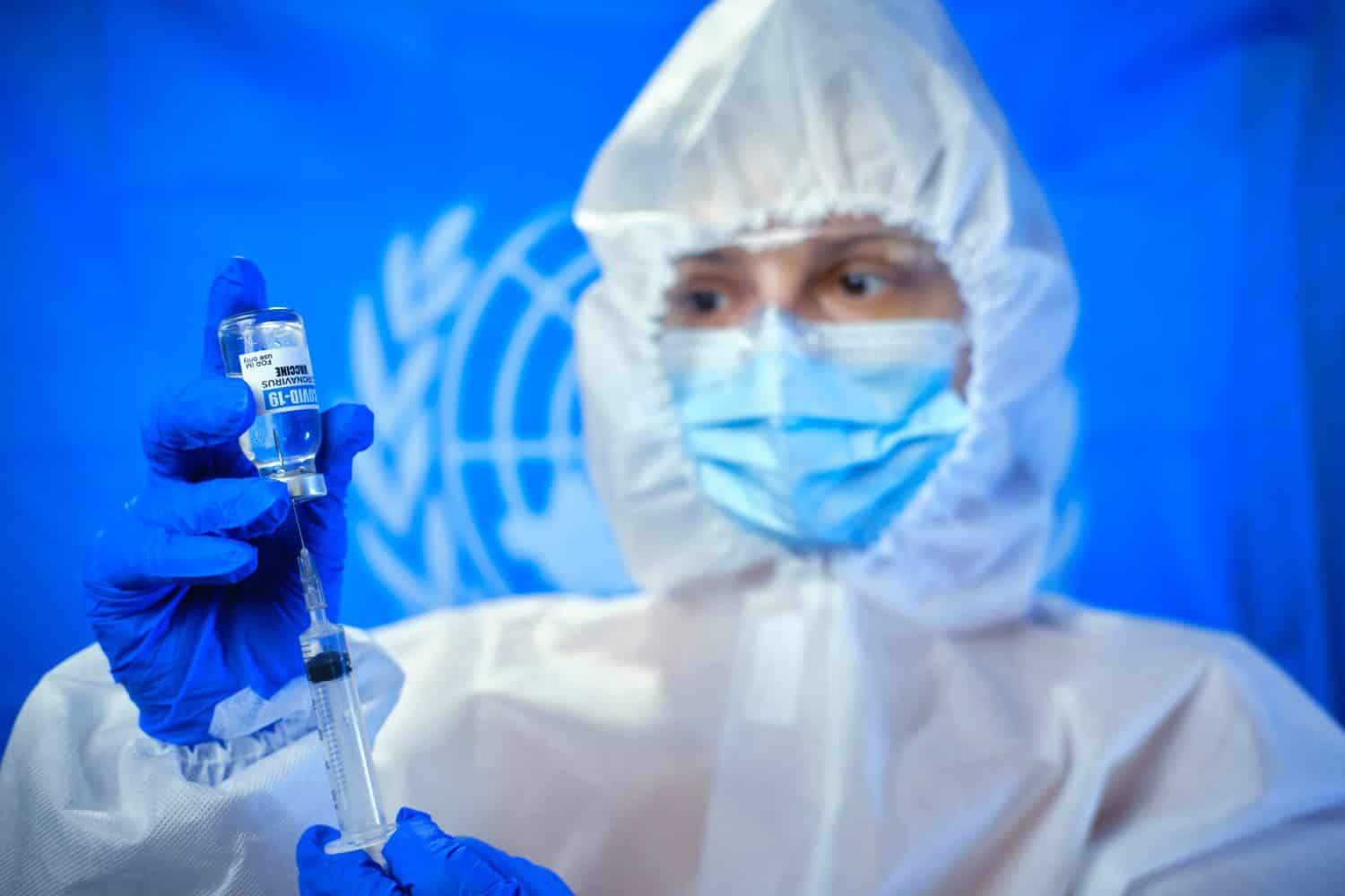 Blured doctor or nurse wearing personal protective equipment and holding syringe of corona covid-19 vaccine in laboratory or hospital against the background of the UN flag